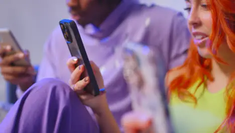 Close Up Studio Shot Of Young Gen Z Friends Sitting On Sofa Talking And Sharing Social Media Post On Mobile Phones 9