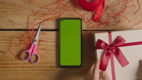 Overhead Shot Of Man Gift Wrapping Romantic Valentines Present Of Perfume In Box Next To Green Screen Mobile Phone