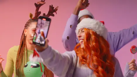 Studio Shot Of Gen Z Friends Dancing And Posing For Selfie At Christmas Party Wearing Santa Hat And Reindeer Antlers 2