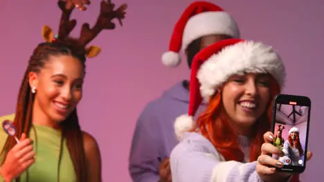 Studio Shot Of Gen Z Friends Dancing And Posing For Selfie At Christmas Party Wearing Santa Hat And Reindeer Antlers