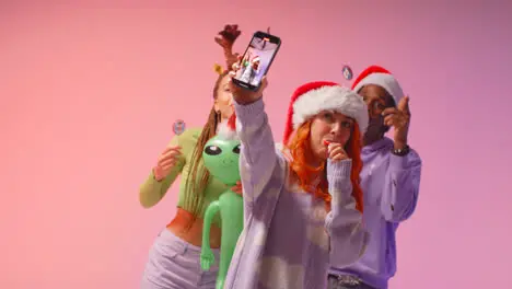 Studio Shot Of Gen Z Friends Dancing And Posing For Selfie At Christmas Party Wearing Santa Hat And Reindeer Antlers 1