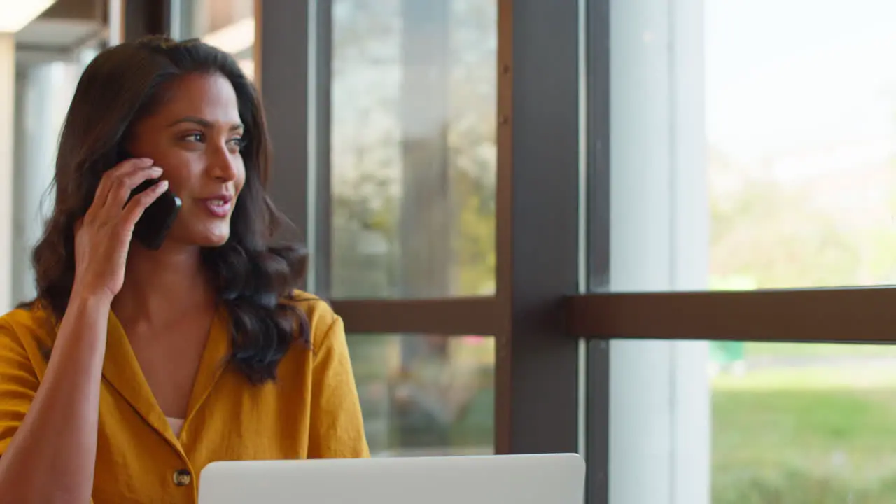 Mature Businesswoman Working On Laptop At Desk In Office Taking Call On Mobile Phone