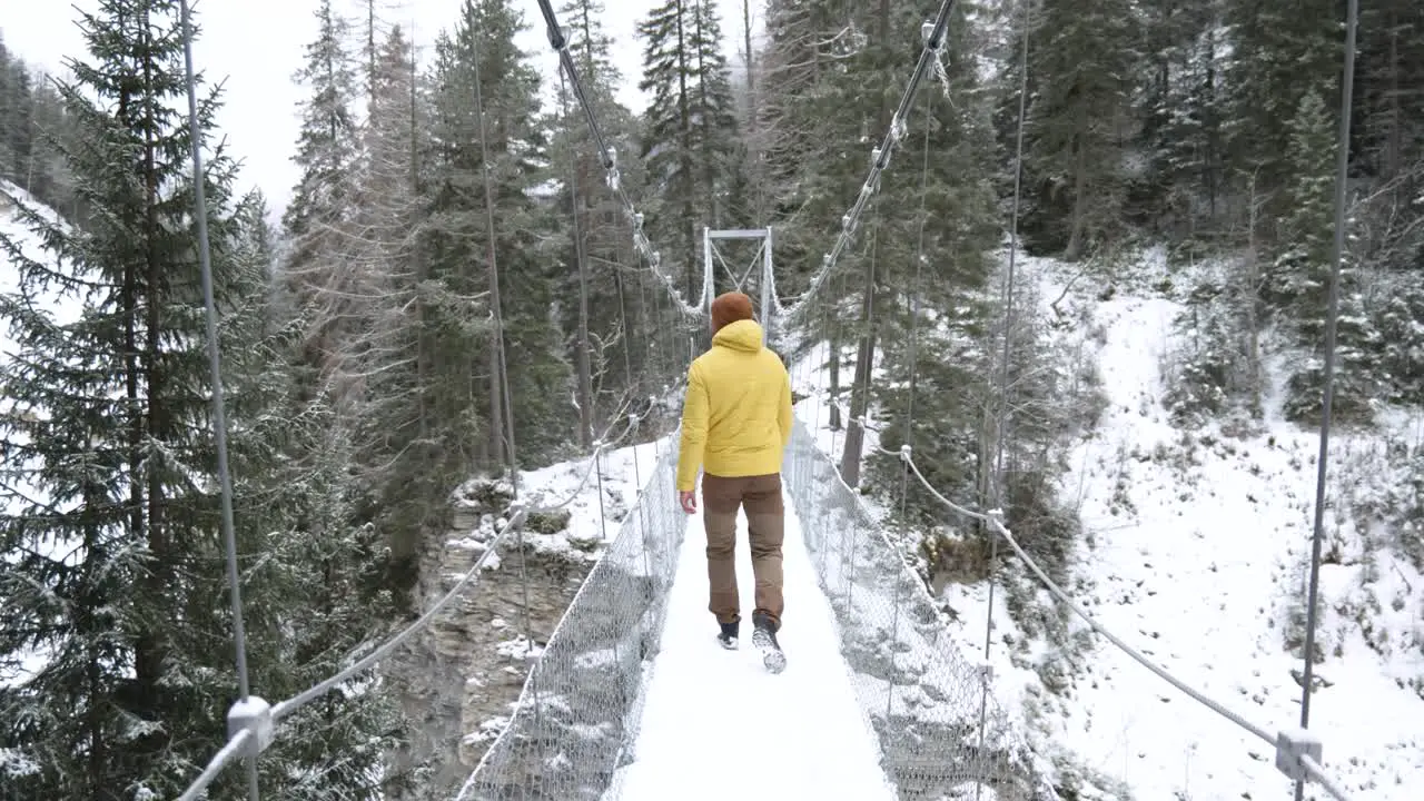 Young caucasian man in yellow winter jacket walks across a hanging bridge in a forest during winter while snowing