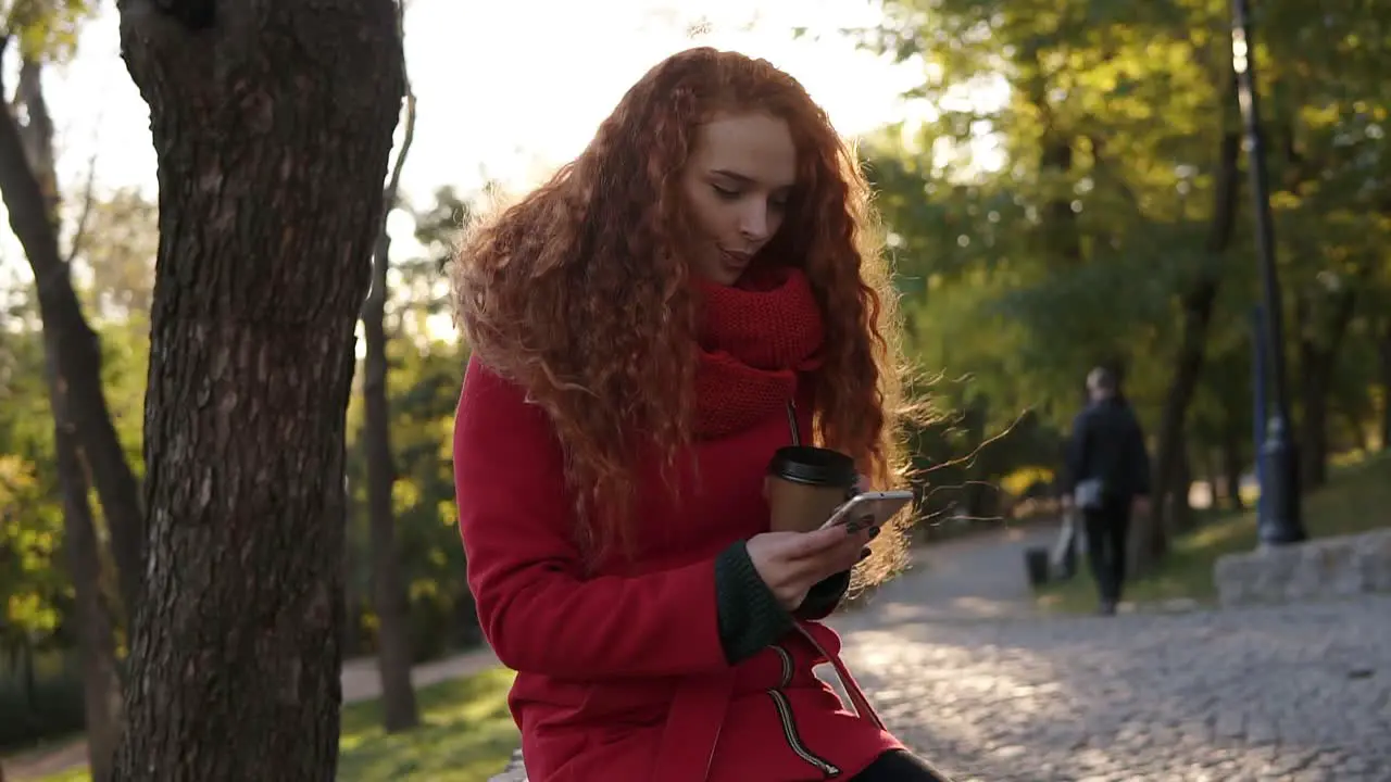 Woman With Curly Long Red Hair In A Red Coat And Scarf Standing In The City Park Using Cell Phone And Drinking Coffee From Corton Park