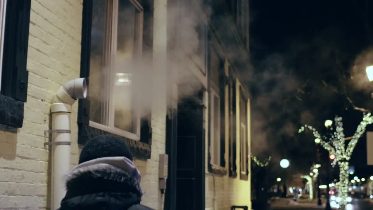 Hooded man walks past a pipe spewing steam and fog out into the cold nighttime air in slow motion