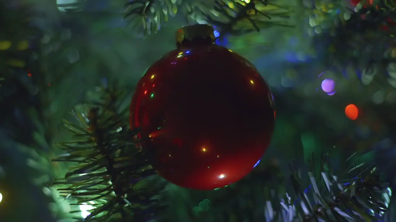 Closeup of dark plain red Christmas tree ornament hanging motionless while multicolored lights blink around it