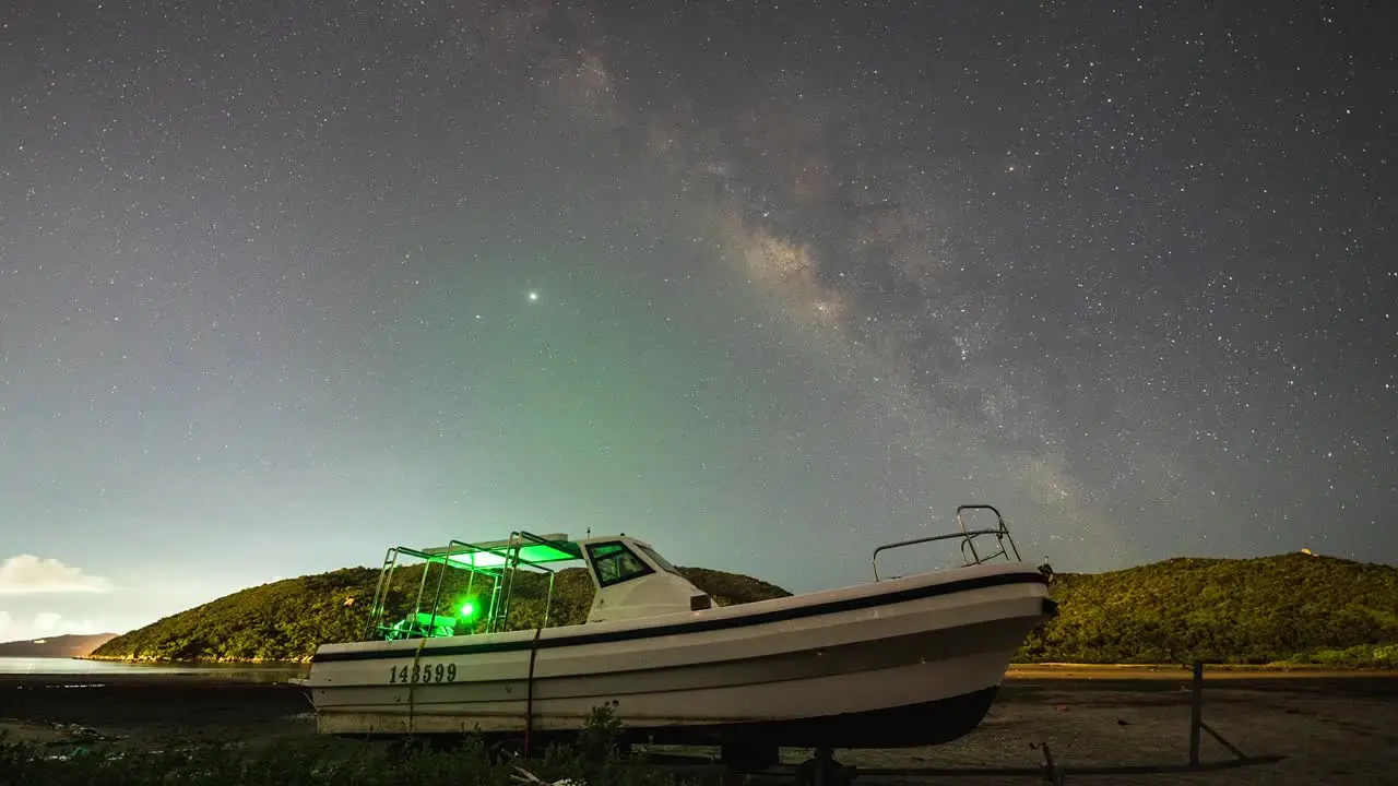 Small empty boat illuminated under Lantau island milky way night sky shooting lights timelapse