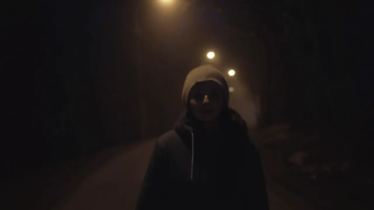 Girl walking dark road in light of street lamps face front view looking into camera