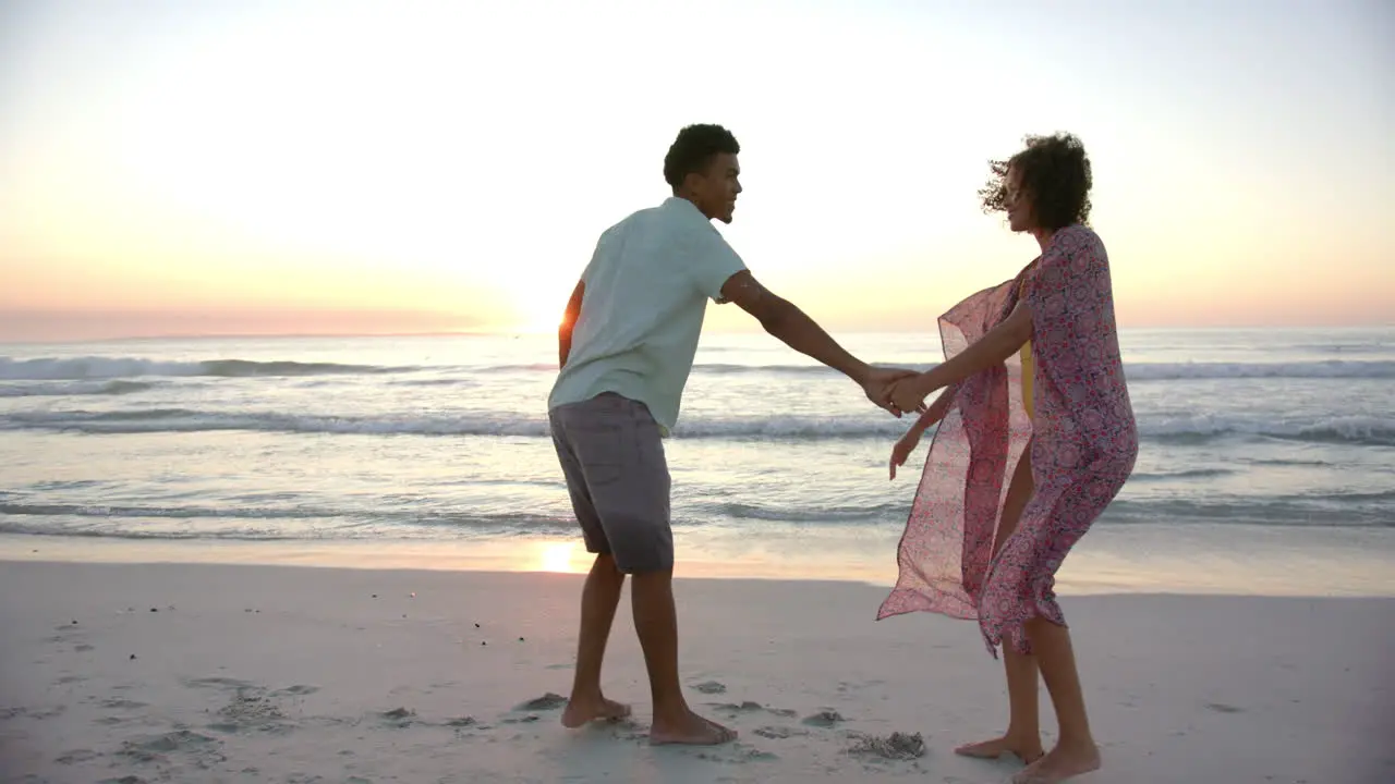 Biracial couple holds hands on a beach at sunset waves gently breaking in the background