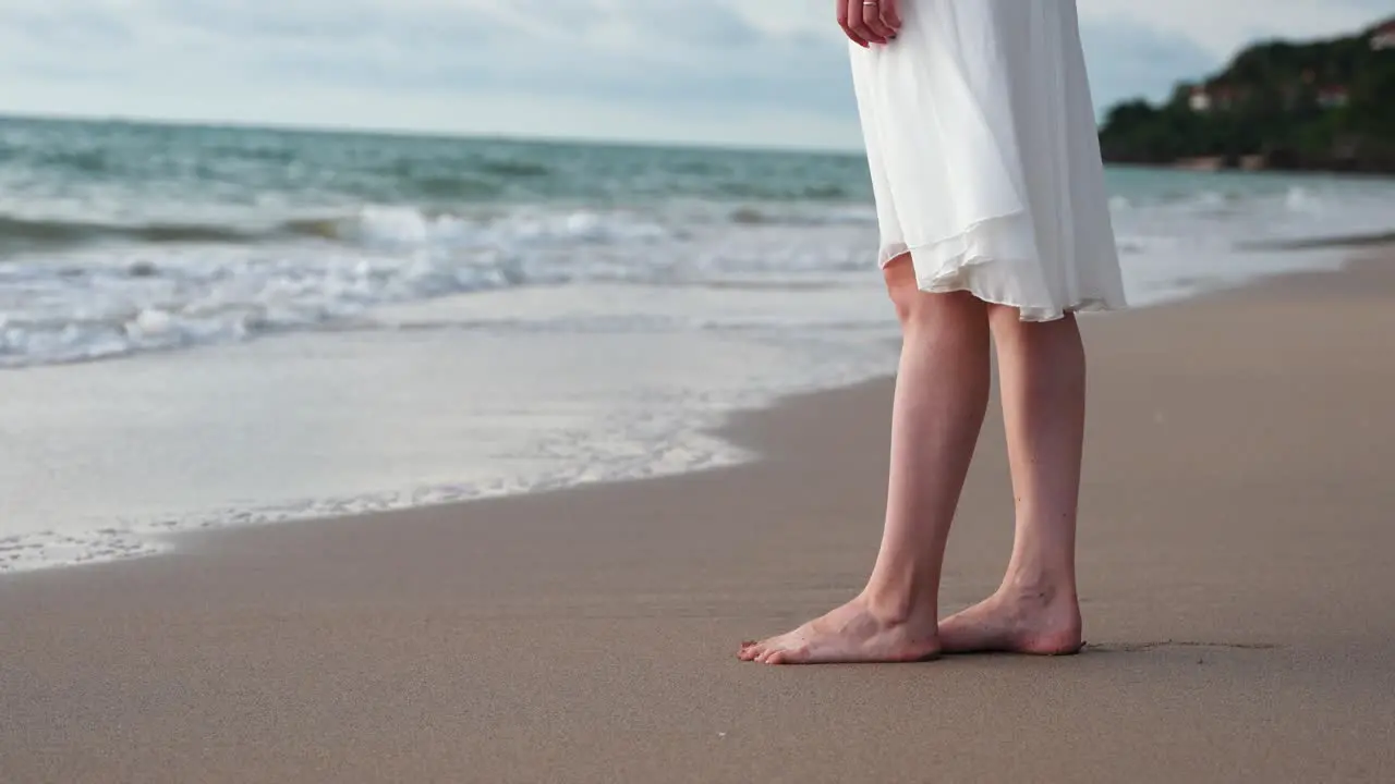 Close up of young woman in white dress bare feet on the beach waves gently touching the shore in slow motion