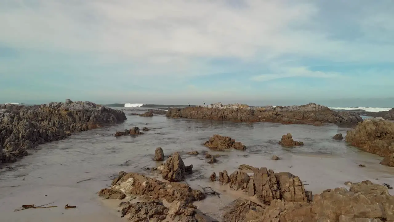 slow and Low flying drone shot from the beach through boulders and birds sitting on rocks rising to reveal open ocean filled with bamboo and waves rolling in partly cloudy conditions