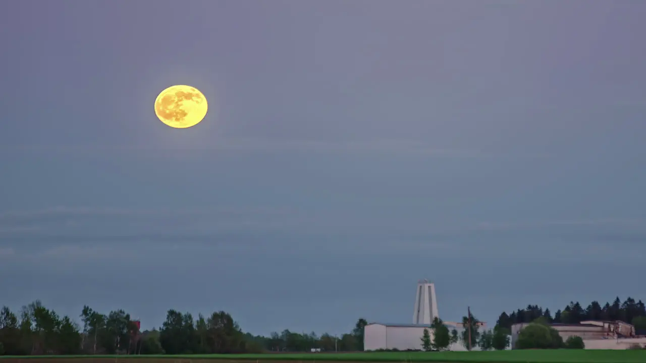 Timelapse video of moon rising over a blue sky in a rural landscape with view of a white building in a distance