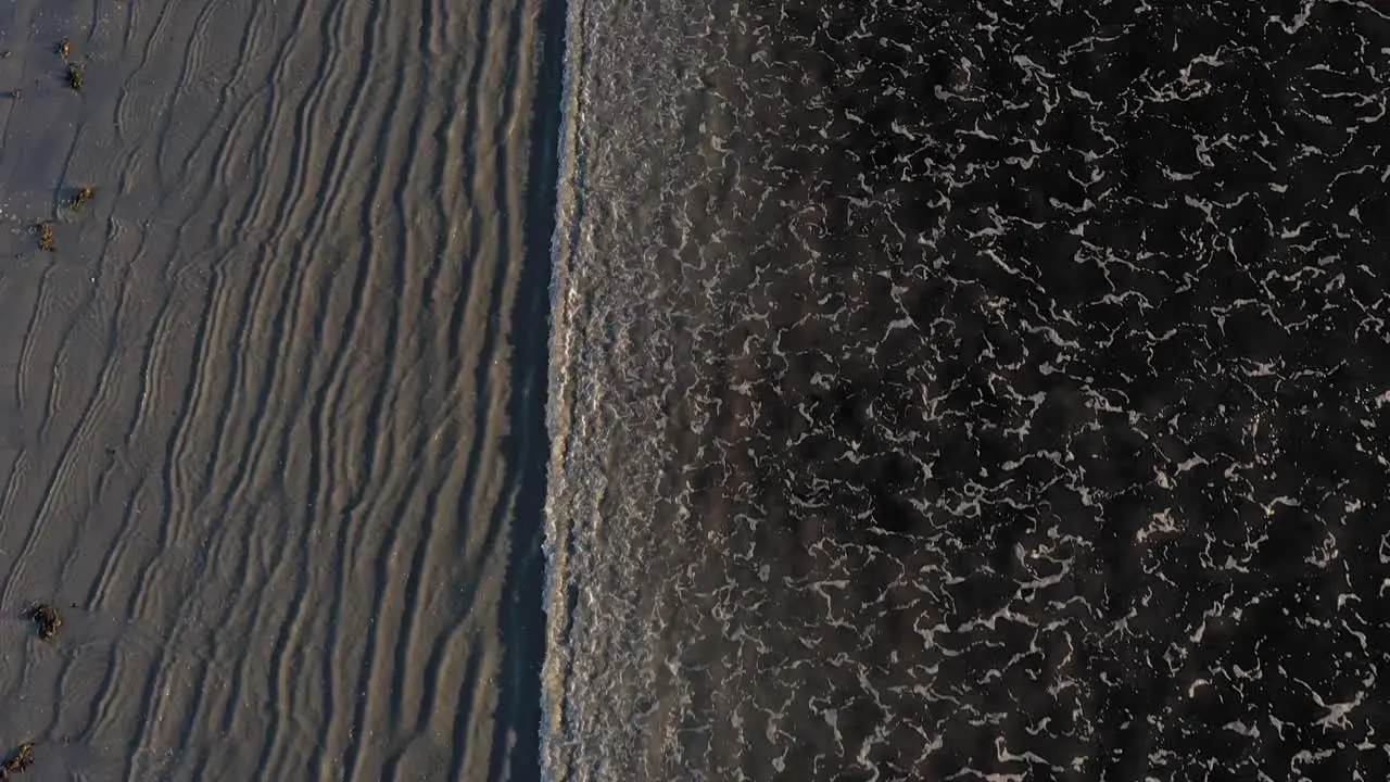 Aerial TOP DOWN flying over a series of waves coming in on a grey sand beach with cool patterns in the sand and surf