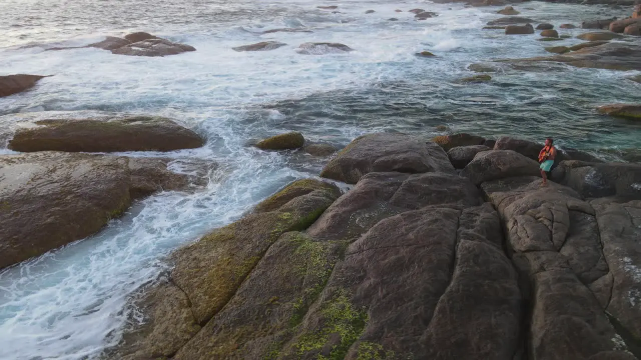 4K Amazing aerial shot attractive young male playing guitar on rocks front of wild sea waves at sunset lifestyle Slow motion