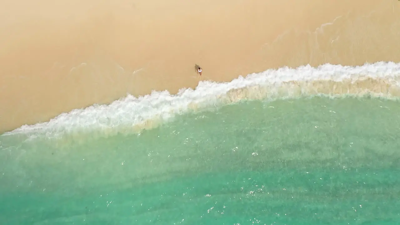 Aerial view of woman in white swimsuit walking on sandy beach into clear turquoise water at tropical island in Indonesia