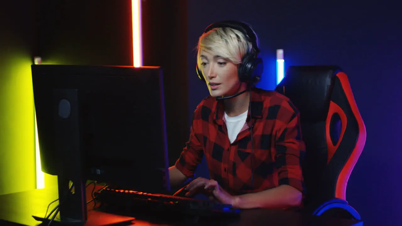 Young Woman With Short Blond Hair In Big Headphones Sitting In The Gamer Chair Playing A Game On The Computer In A Room With Colorful Neon Lamps On The Wall