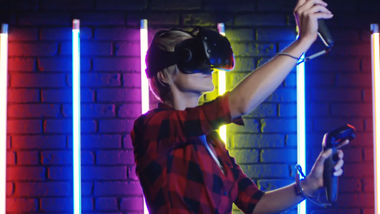 Young Woman In Vr Glasses And Using Joystick While Playing A Virtual Reality Game In A Room With Colorful Neon Lamps On The Wall 4