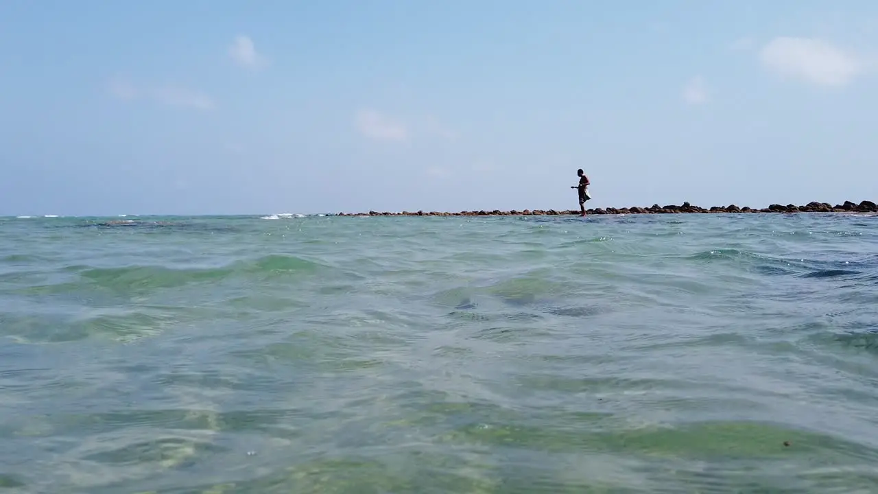 From under water to above water as a fisherman stands alone on a rock at high tide slowly reeling his line in