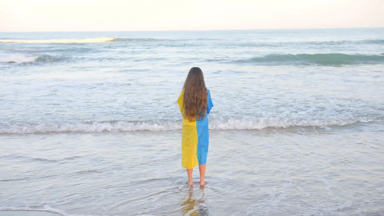 Young Girl With Curly Blonde Hair Carrying A Ukraine Flag On Her Back Watching Sea Waves Rolling To The Shore