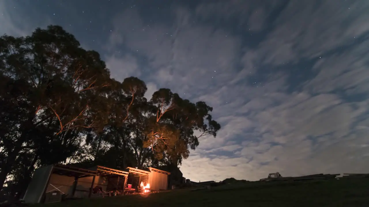 A zoomed in time lapse of a man having a camp fire by a shed at night under some gum trees with clouds rolling past and the night time stars in the background