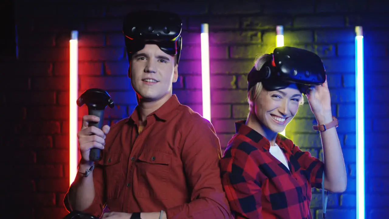 Young Couple Of Friends Man And Woman Taking Off Vr Glasses After A Game Set And Smiling To The Camera In A Room With Colorful Neon Lamps On The Wall