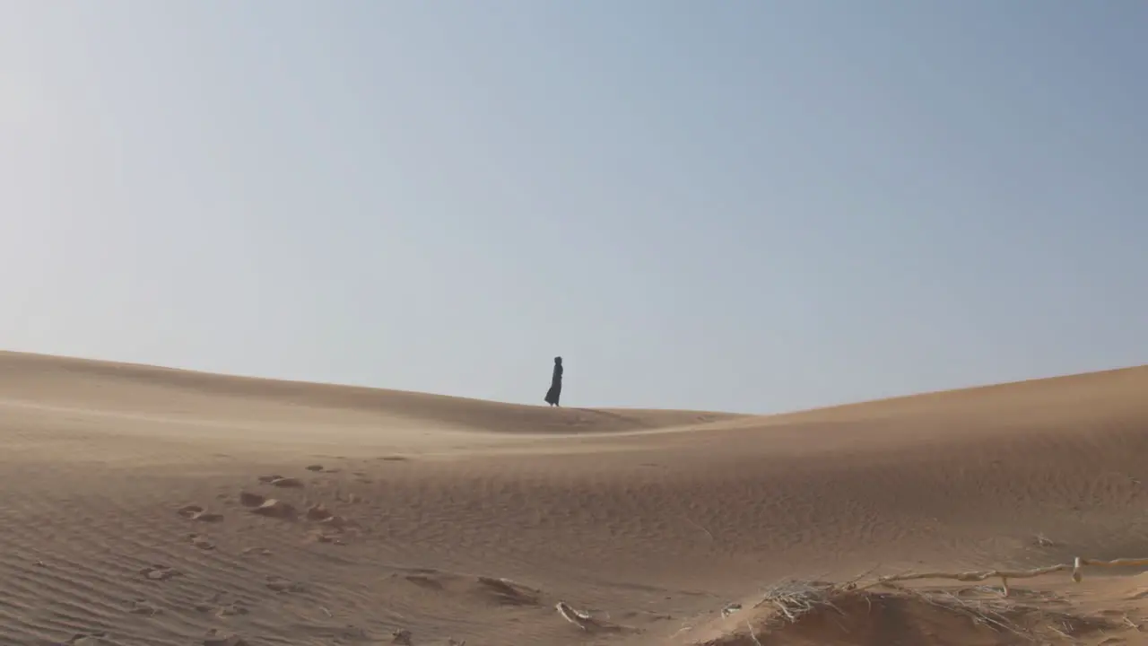 Extreme Long Shot Of A Muslim Woman In Traditional Dress And Hijab Walking In A Windy Desert