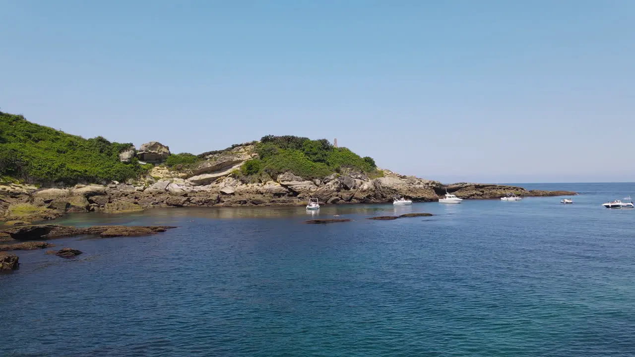 Zoom In Shot Of Blue Sea With Rocky Coast While Some Yachts Are Anchored Near The Shore And People Swimming