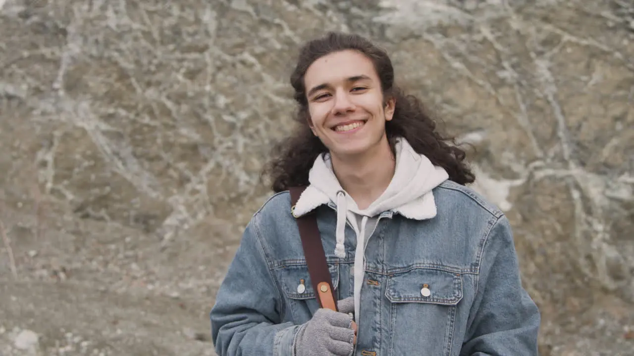 Front View Of A Teenage Boy With Long Hair And Winter Clothes Smiling And Looking At Camera On The Mountain On A Windy Day