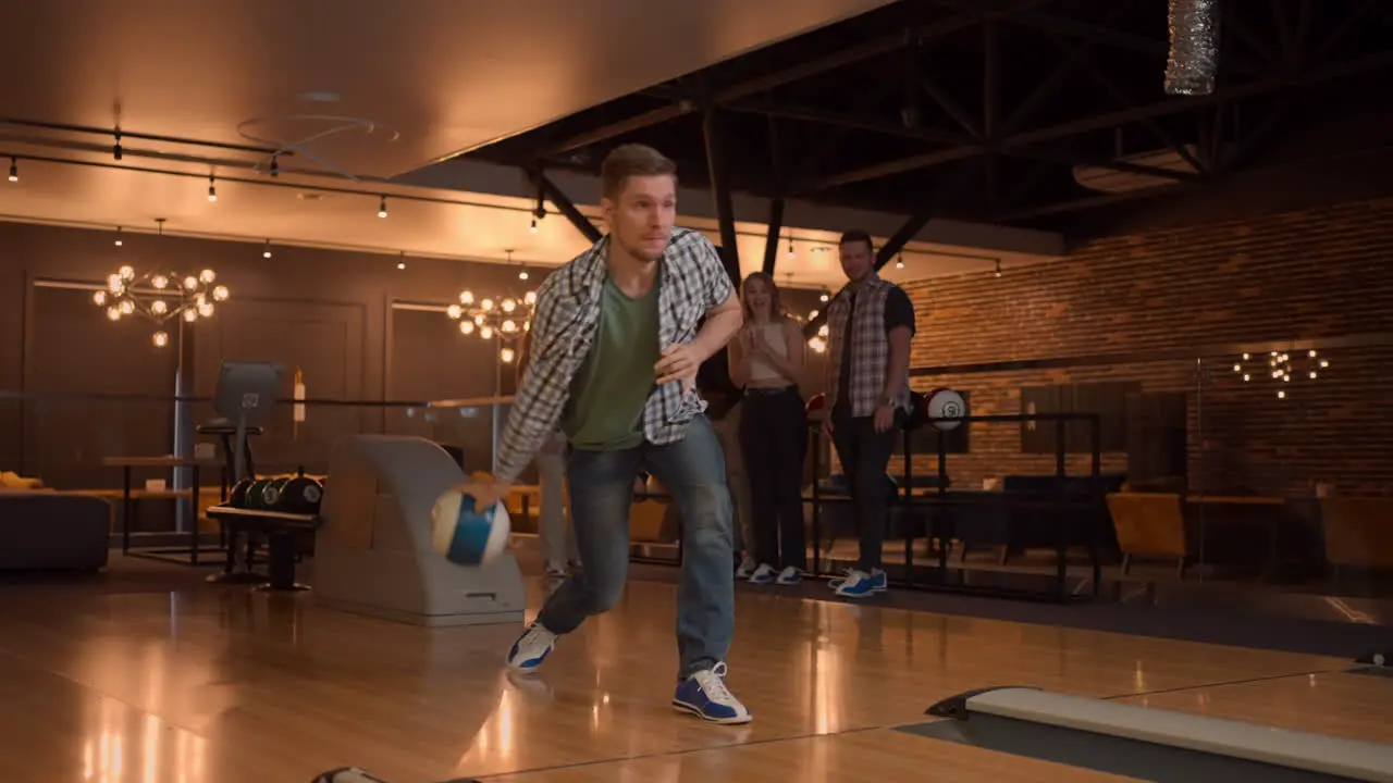 A man in a bowling alley throws a ball on the track and knocks out a shot in slow motion and jumps and dances for joy A group of multi-ethnic friends play bowling together clapping happy supporting