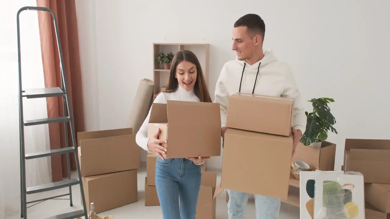 Happy Couple Moving To New Place Holding Cardboard Boxes And Walking Together In Empty Flat