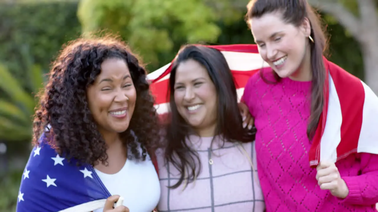 Three happy diverse female friends standing with flag on backs in sunny garden