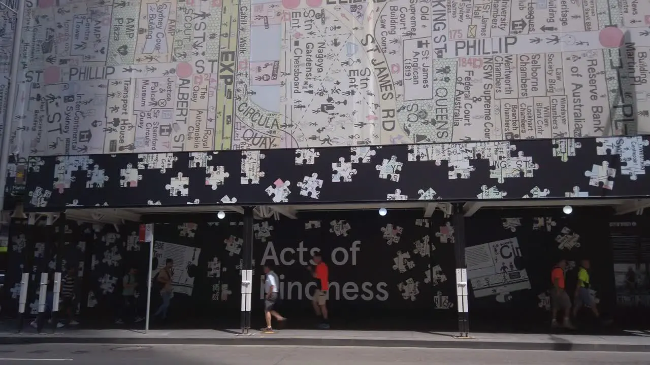 People walking on a sidewalk in Sydney CBD downtown shopping district with the Act of Kindness writing with map of Sydney city building art above them