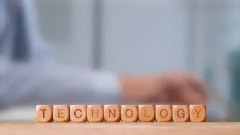 Business Concept Wooden Letter Cubes Or Dice Spelling Technology With Person Working On Laptop In Office In Background