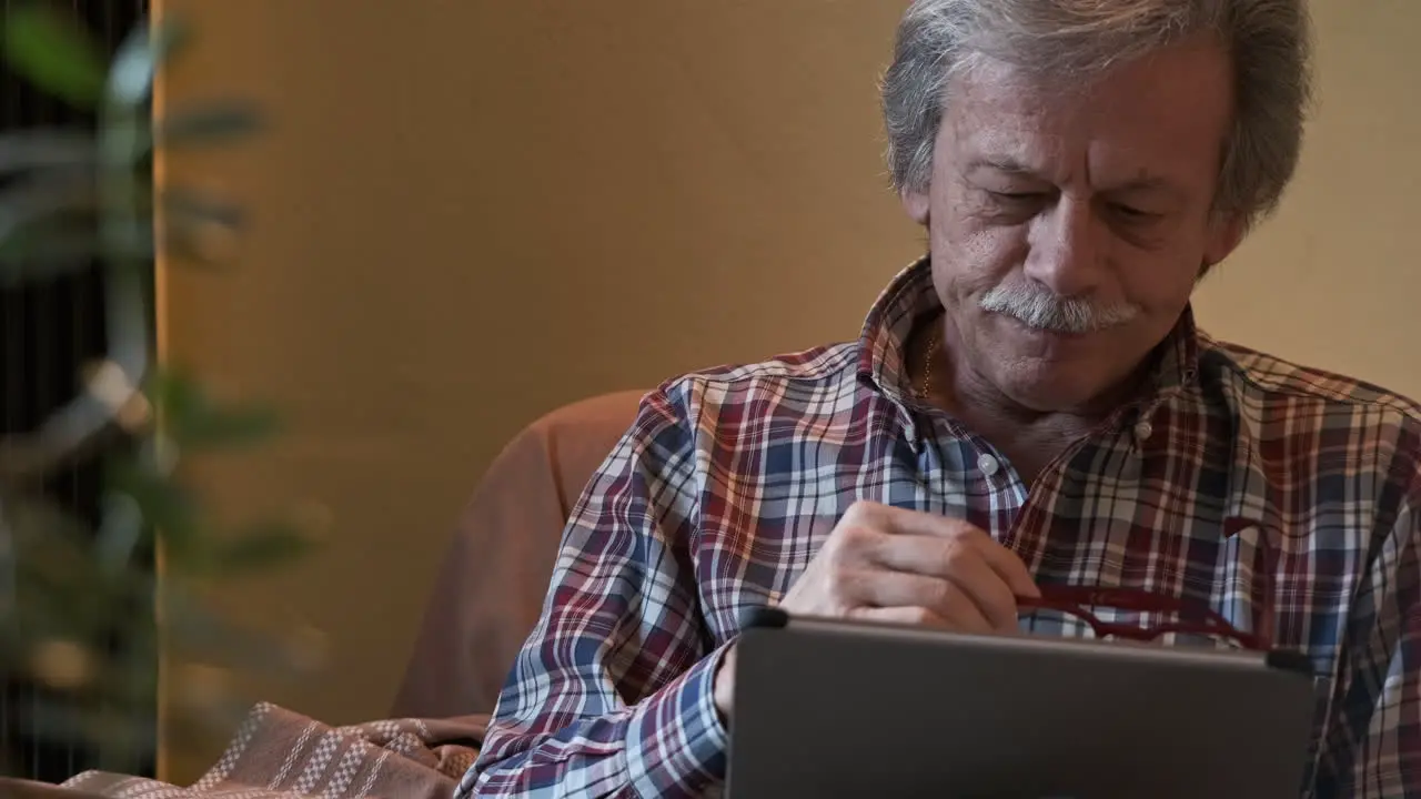 Front View Of A Man Sitting On Couch Using Tablet With Gray-Haired Takes Off His Glasses And Looks In Front Slow Motion Shot