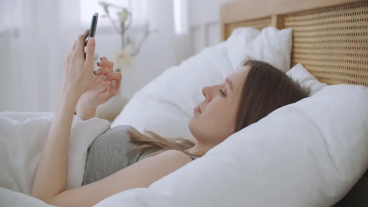 Woman in morning sitting in bedroom leaning against the soft leather back of bed Write messages with your smartphone use apps to information