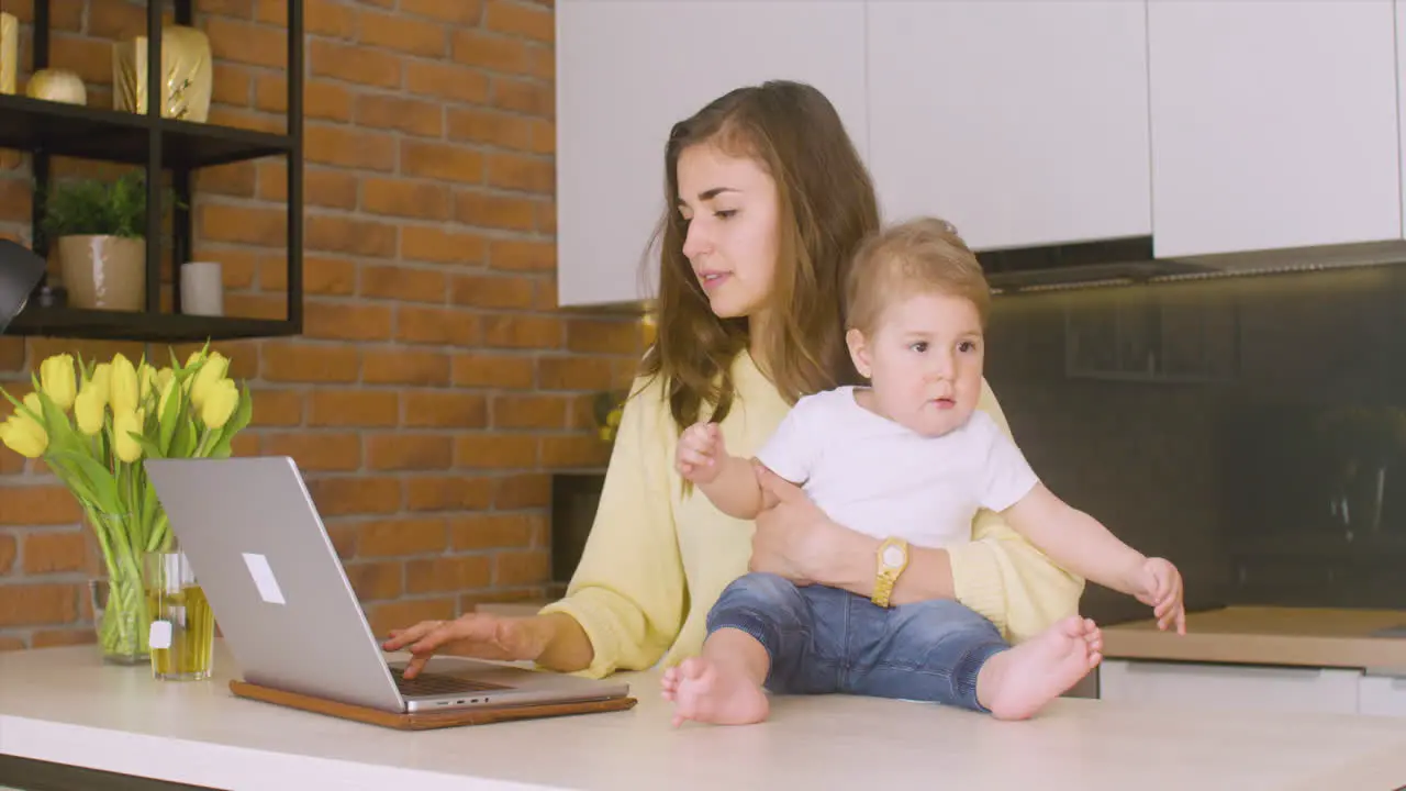 Woman Holding His Baby Sitting On The Kitchen Counter While Using The Laptop