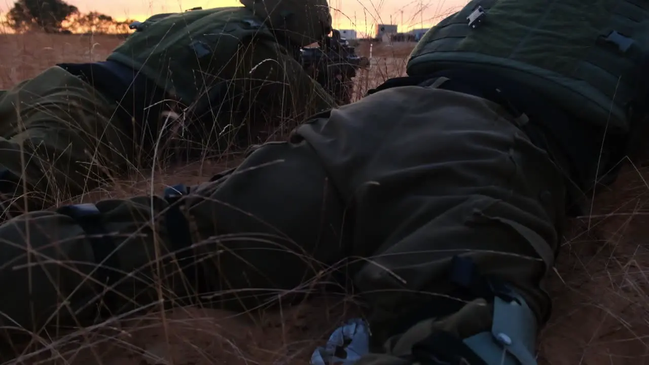 Two combat soldiers lie on the sandy desert ground pointing their weapons at the buildings on the horizon