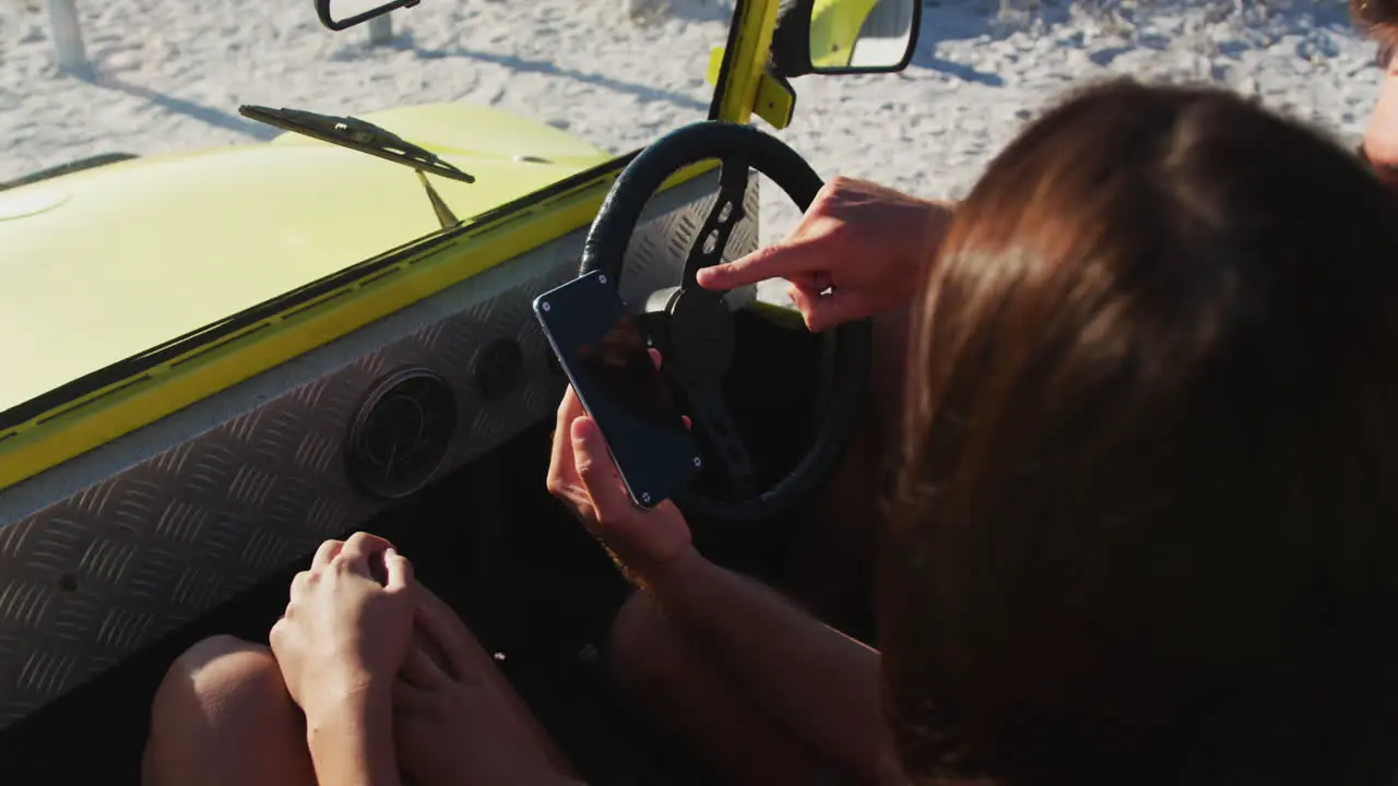 Happy caucasian couple sitting in beach buggy by the sea using smartphone