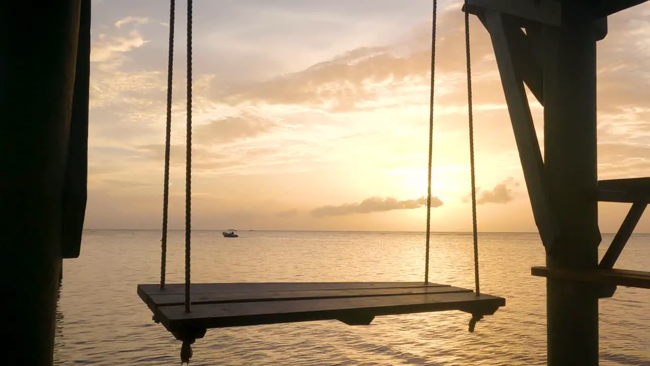 An empty wooden swing is swinging on a dock overlooking the ocean with a boat in the orange rays of the sunset