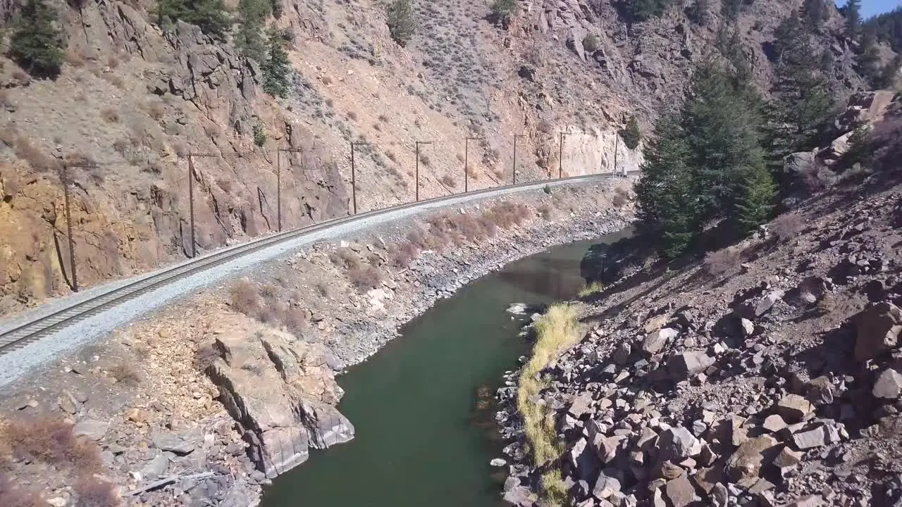 trucking aerial shot of a railroad track following an emerald green river with power lines and pine trees in a western setting with lots of rocky terrain and hills