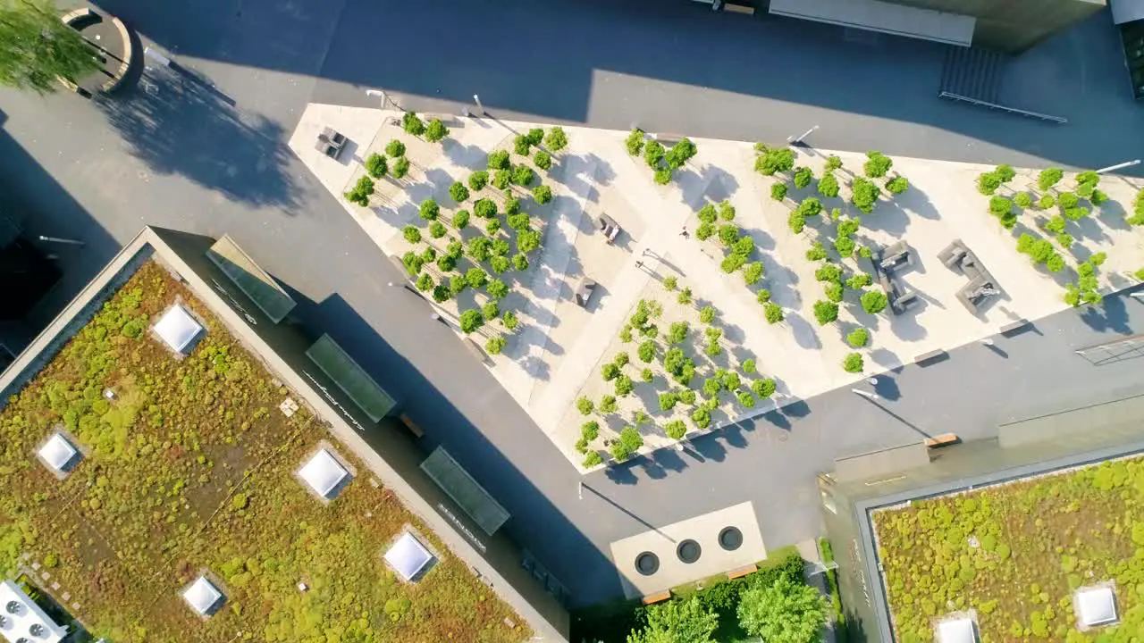 Aerial birdseye view of a modern plaza with angular shapes small green trees people and surrounded by shops with greed roofs pull back