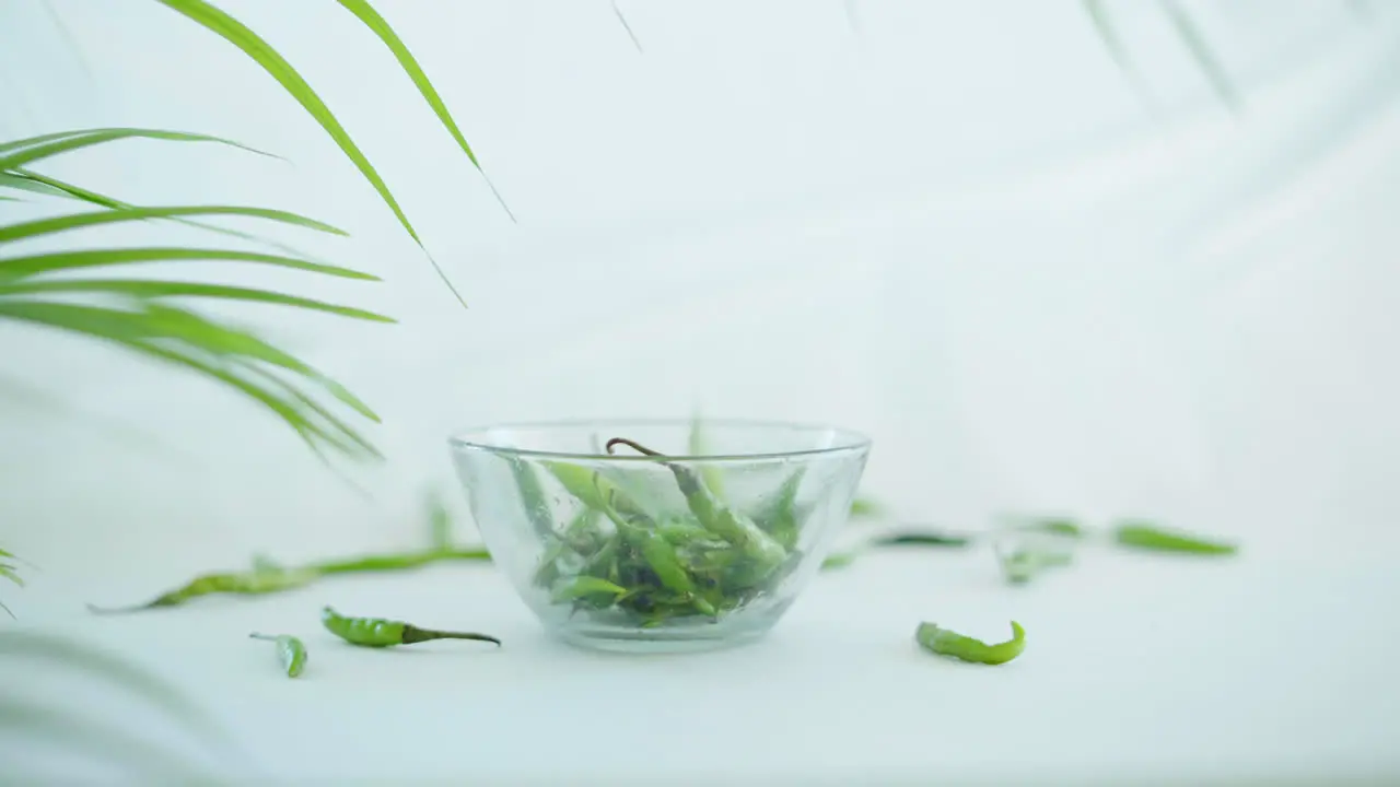 Zoom out shot of green chillis slowly falling in to a glass bowl on a white background