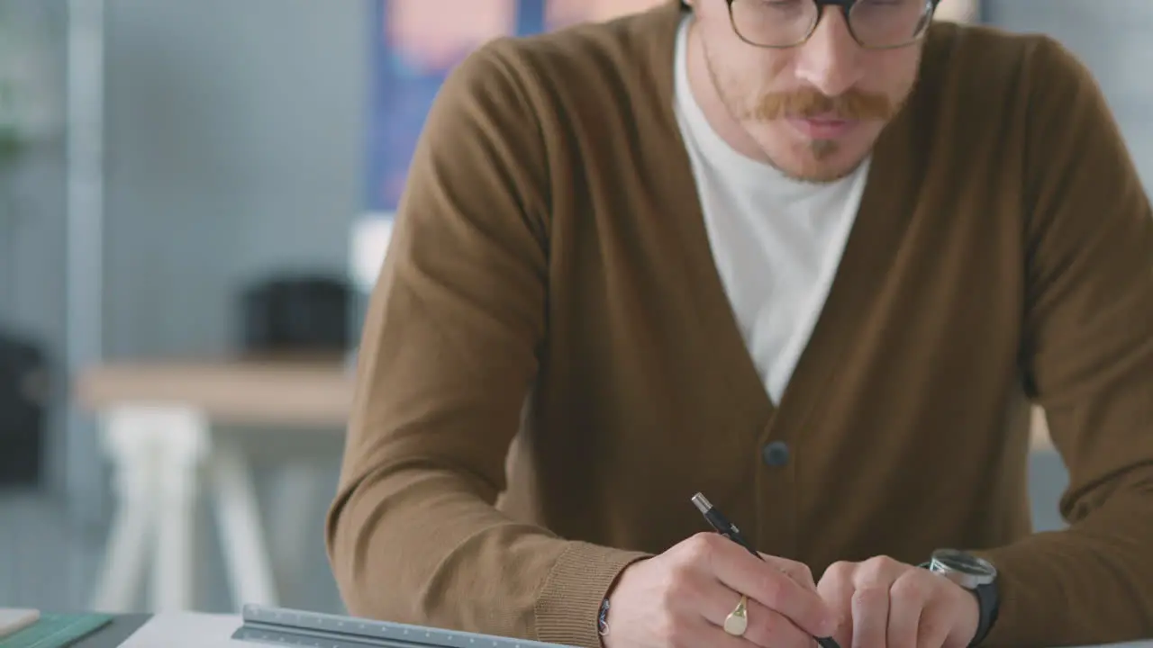 Close Up Of Male Architect In Office Working At Desk With Model Of Building Making Notes On Plan