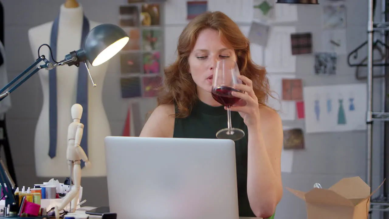 Female Owner Of Fashion Business With Glass of Wine Working Late On Laptop In Studio