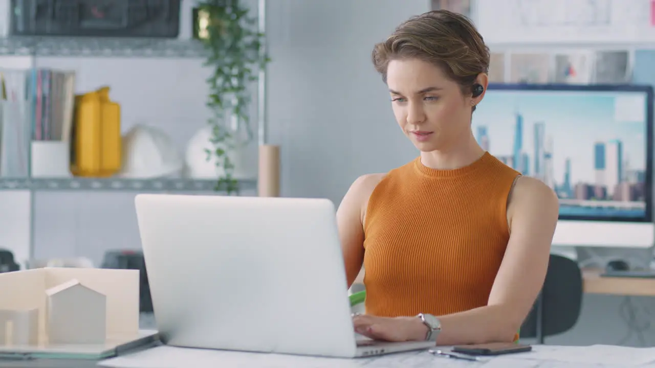 Female Architect In Office Listening To Music On Wireless Earphones Whilst Using Laptop