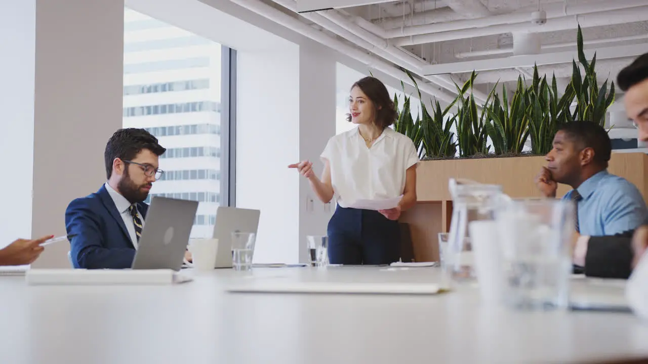 Businesswoman Standing Giving Presentation To Colleagues Sitting Around Table In Modern Office