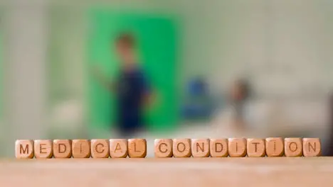 Medical Concept With Wooden Letter Cubes Or Dice Spelling Medical Condition Against Background Of Nurse Talking To Patient In Hospital Bed