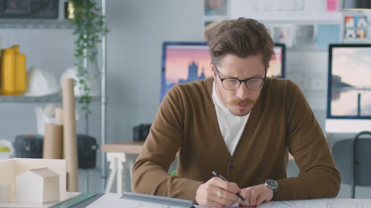 Male Architect In Office Working At Desk With Model Of Building Making Notes On Plan