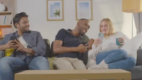 Multi-Cultural Friends Sitting On Sofa At Home Together All Looking At Their Mobile Phones