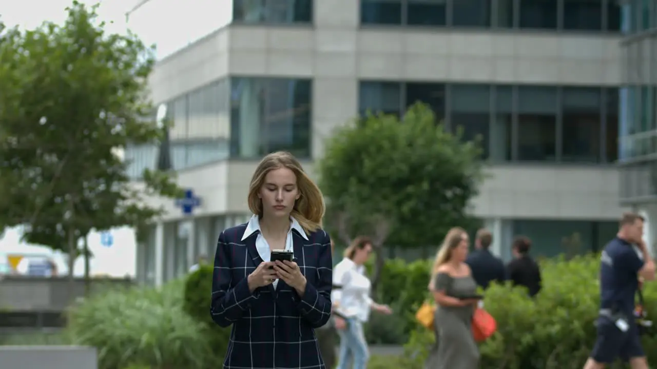 In a clean urban setting a young women walks towards the camera paying extreme attention to her cell phone